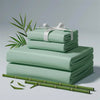 A stack of light green bamboo bed sheets rolled and tied with a white ribbon, resting on a surface with green leaves.