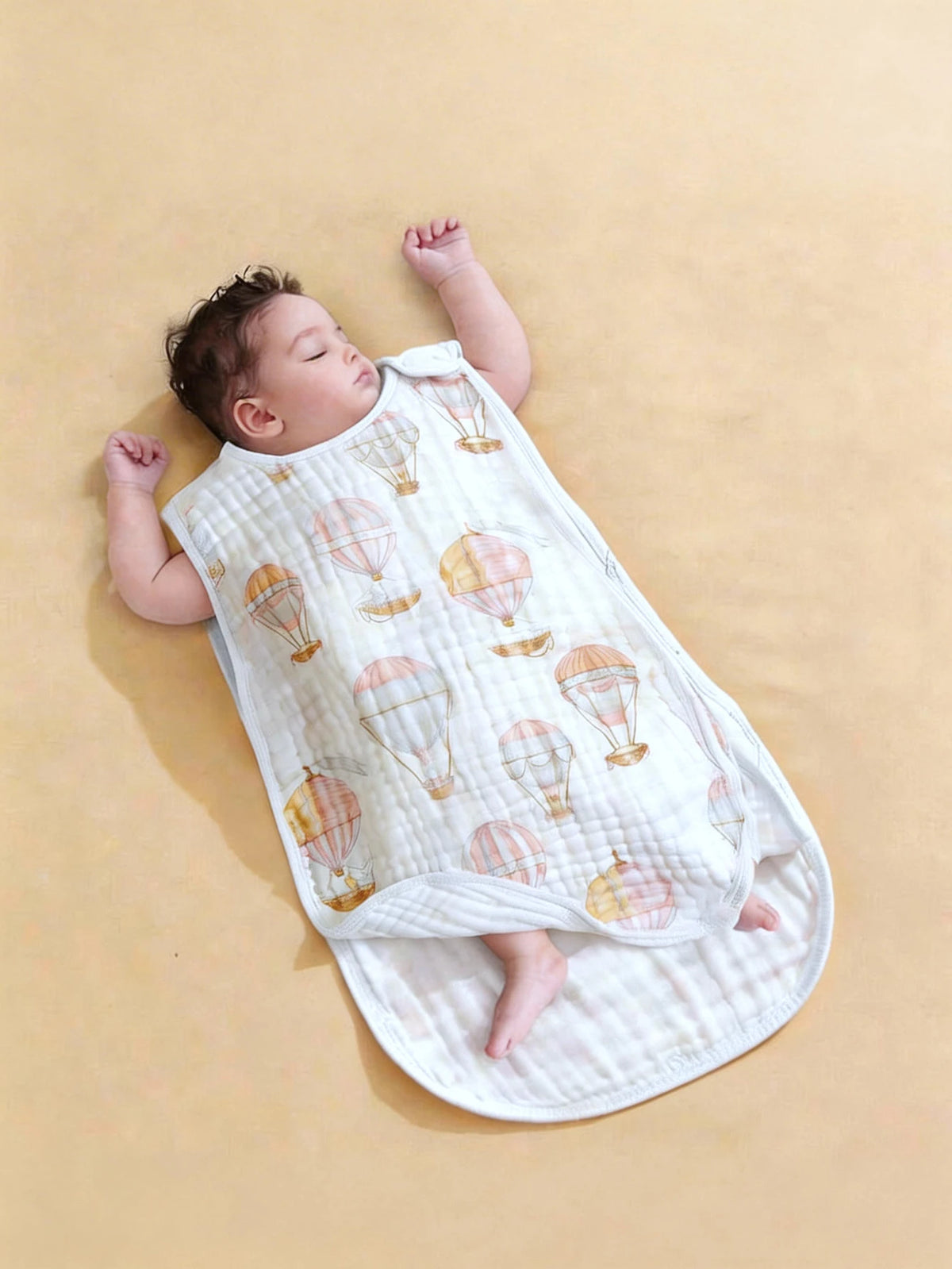 A baby is sleeping on a white fabric sleeping bag patterned with orange and gold hot air balloons.