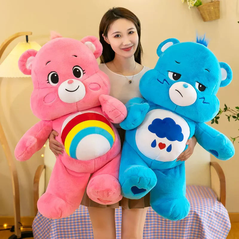 A woman is holding a pink Care Bear Rainbow Bear plush toy and a blue Care Bear plush toy.