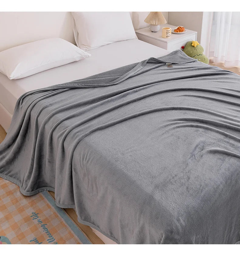 A gray extra soft flannel throw blanket draped over a white bed with a green stuffed animal nearby.