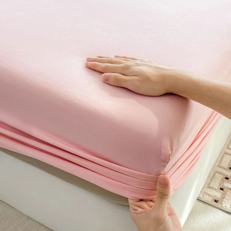 A hand is folding a pink fitted bed sheet made of premium 100% cotton.