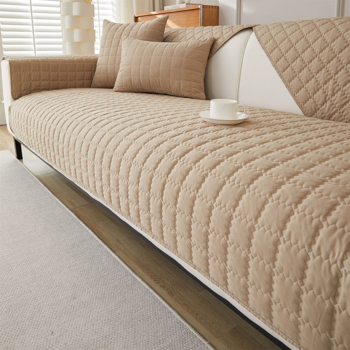A beige quilted anti-slip sofa cover featuring a plush texture on a white sofa with neutral pillows.