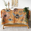 A bohemian geometric throw blanket featuring floral patterns, eyes, and a bee design draped over a wooden bench.