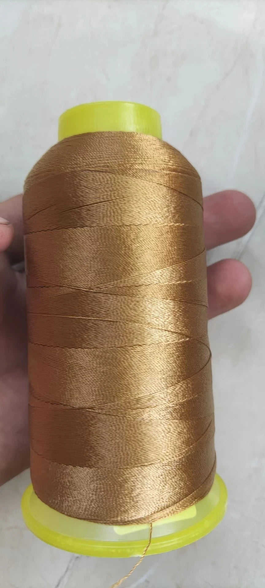 A spool of golden brown high tenacity polyester sewing thread with a yellow plastic core is held in a hand.