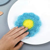 A hand is placing a bright blue and yellow fluffy kitchen sponge onto a white plate.
