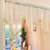A sheer, cream-colored curtain with gold lace beading and tassels hangs in a window frame.