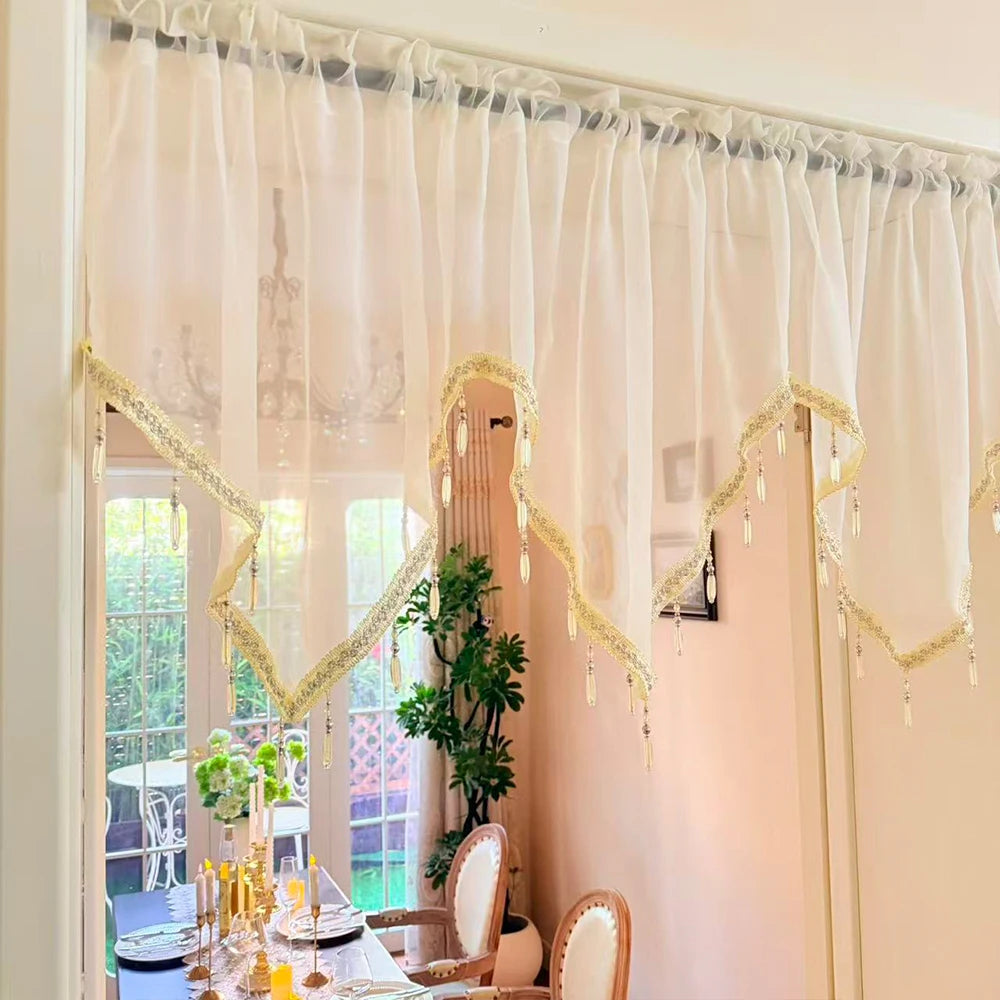 A sheer, cream-colored curtain with gold lace beading and tassels hangs in a window frame.