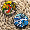 A pair of metal broach pins featuring the anime characters Ho-Oh and Lugia set against a woven straw background.