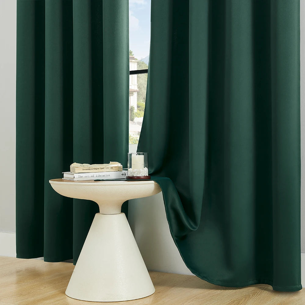 A solid dark green blackout window curtain hanging open, revealing a white pedestal with books and a glass.