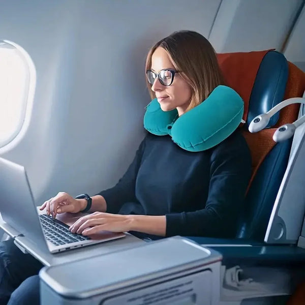 A woman wearing glasses is using a laptop while resting her head on a teal inflatable neck pillow in an airplane seat.