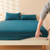 A teal quilted mattress protector cover is displayed on a bed with matching pillows and a hand adjusting the fabric.
