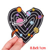 A black heart shaped iron-on patch embroidered with colorful planets and stars.