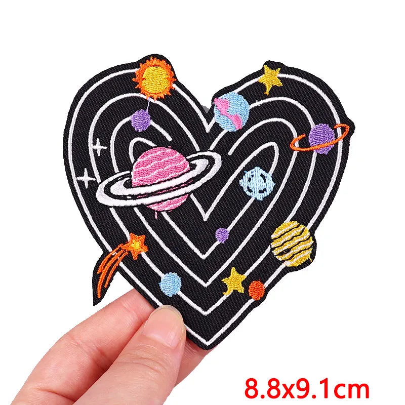 A black heart shaped iron-on patch embroidered with colorful planets and stars.