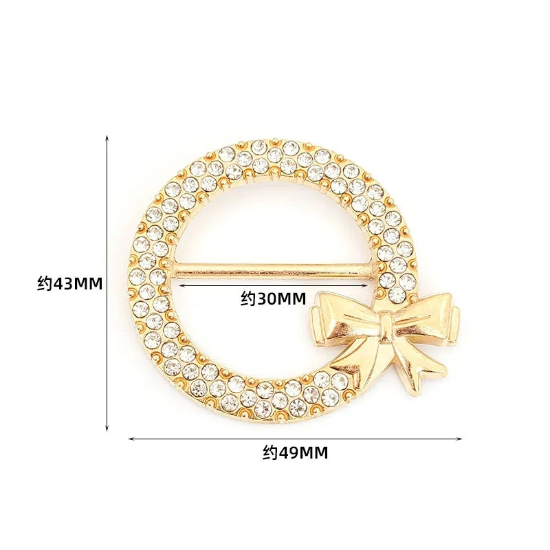 A gold-toned metal ring clip featuring a circular design encrusted with clear stones and accented by a bow detail.