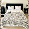 A white leopard print jacquard knit throw blanket is displayed on a bed with white pillows and dark bedding.