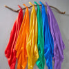 A collection of vibrant silk scarves in red, orange, yellow, green, blue, and purple hanging on a wooden branch.