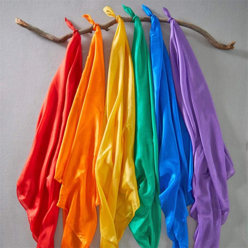 A collection of vibrant silk scarves in red, orange, yellow, green, blue, and purple hanging on a wooden branch.