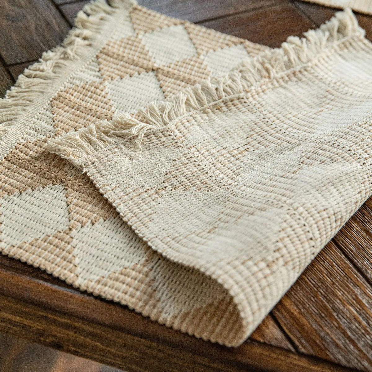 A beige linen table runner with a diamond pattern and frayed edges resting on a dark wooden surface.