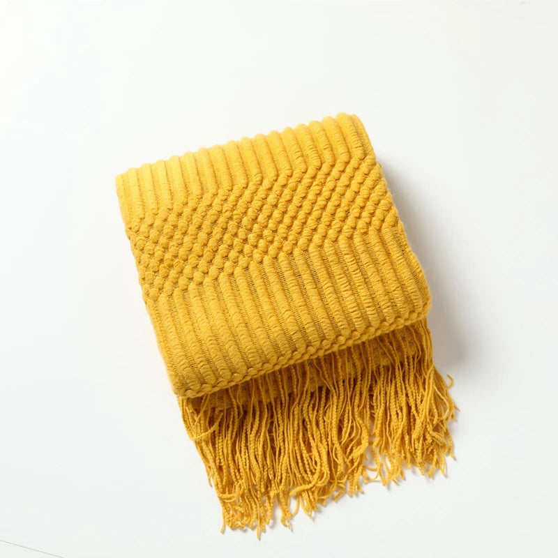 A mustard yellow oversized knitted tassel throw blanket folded and draped on a white surface.
