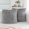 Two gray faux fur throw pillows resting on a white surface next to a window with decorative items.