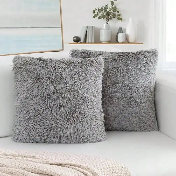 Two gray faux fur throw pillows resting on a white surface next to a window with decorative items.