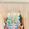 A sheer, beige curtain decorated with gold lace and tassels hangs in a room with a wooden table set for a meal.