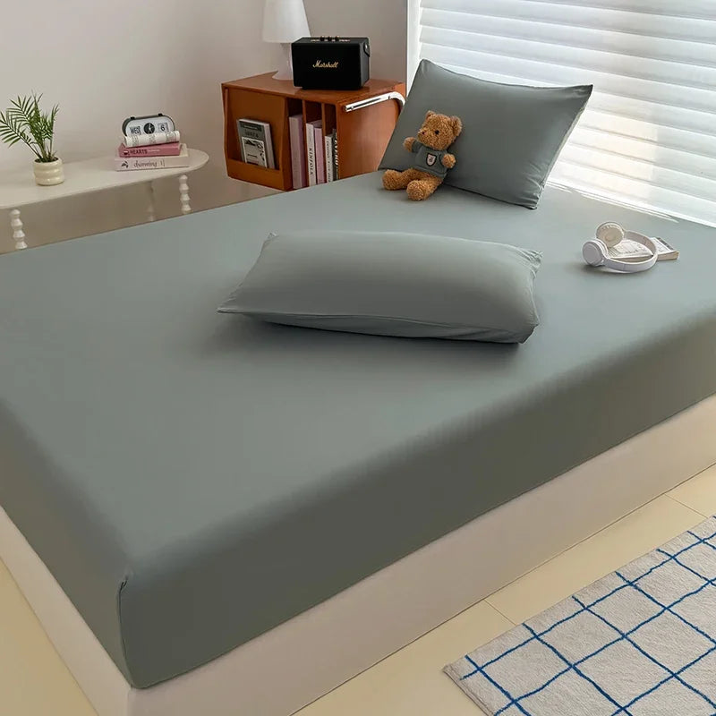 A fitted bed sheet cover in a solid gray color is displayed on a bed, with pillows and a wooden nightstand nearby.