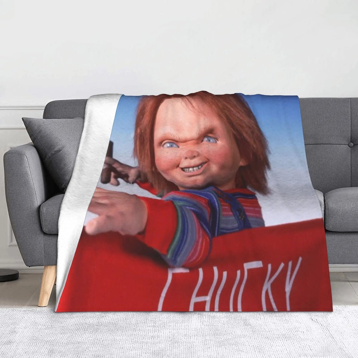 A red flannel throw blanket featuring a graphic of Chucky from the horror movie on a gray sofa.