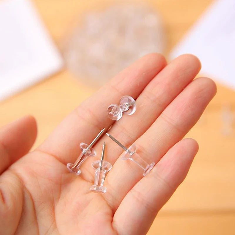 A hand holds several clear plastic push pins with steel tips on a wooden surface.