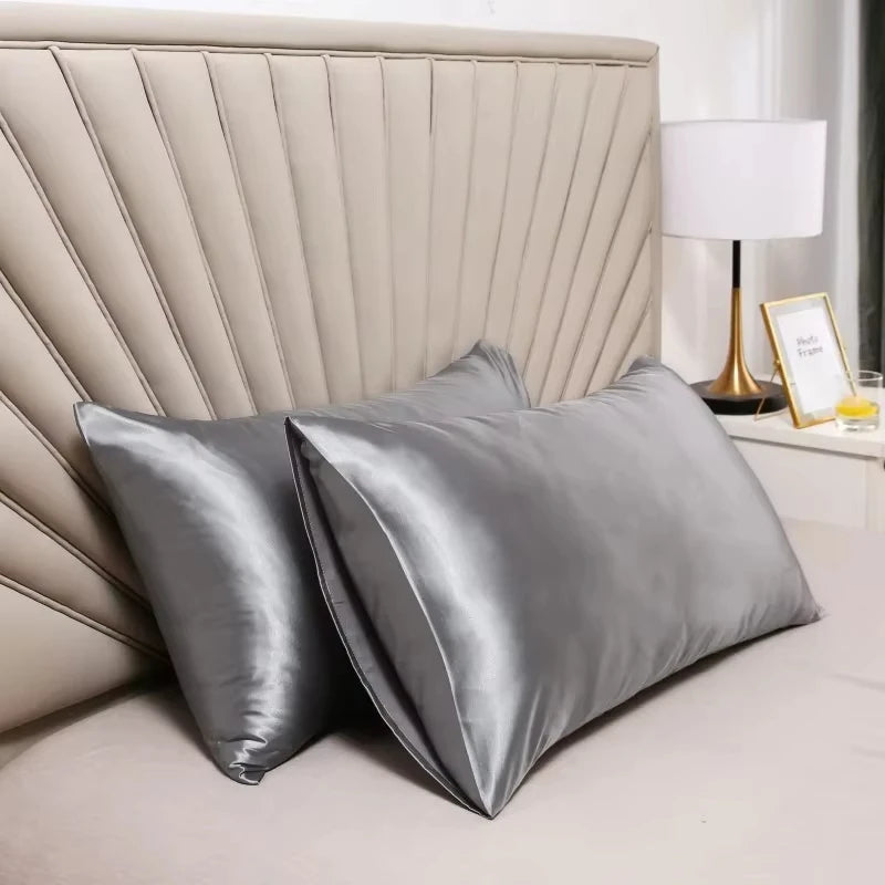A pair of smooth gray satin silk pillow covers resting on a white bed with a padded headboard and a lamp.
