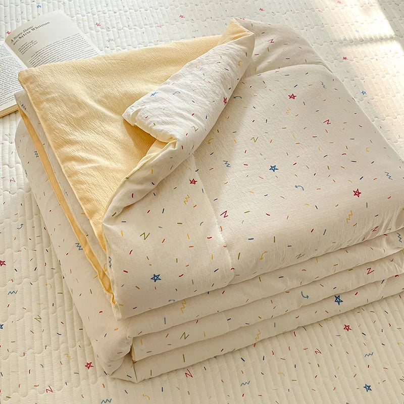 A stack of light beige cartoon quilting comforters and a yellow blanket on a white quilted surface.