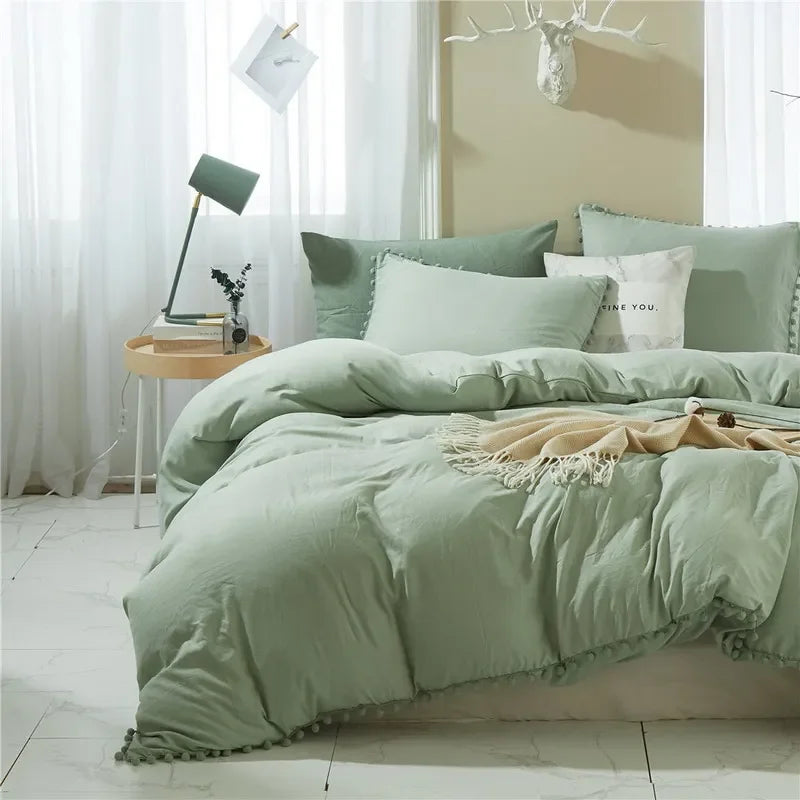 A light sage green duvet cover set with pom-pom decoration displayed on a bed in a bright room.