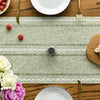 A green embroidered cotton linen table runner with white tassels laid on a wooden table.