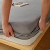 A hand is folding a gray cotton fitted sheet with deep pockets and elastic edges on a wooden bed frame.