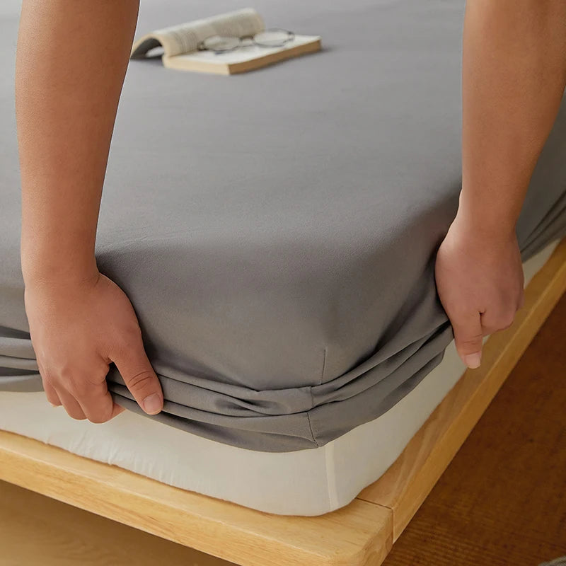 A hand is folding a gray cotton fitted sheet with deep pockets and elastic edges on a wooden bed frame.