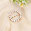 A gold-toned ring clip made of small white pearls, displayed on a soft beige fabric with greenery.