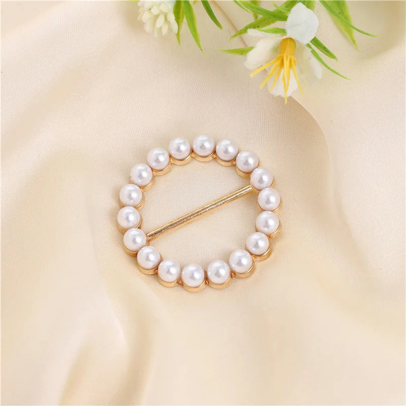 A gold-toned ring clip made of small white pearls, displayed on a soft beige fabric with greenery.