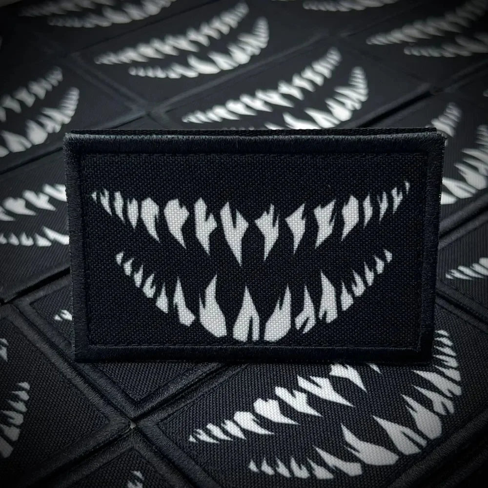A black tactical hook and loop emblem patch is displayed against a dark background with repeating white tooth patterns.