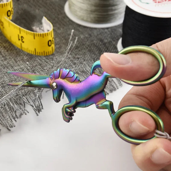 A pair of iridescent, multi-colored stainless steel unicorn sewing scissors held by a hand against a gray fabric background.