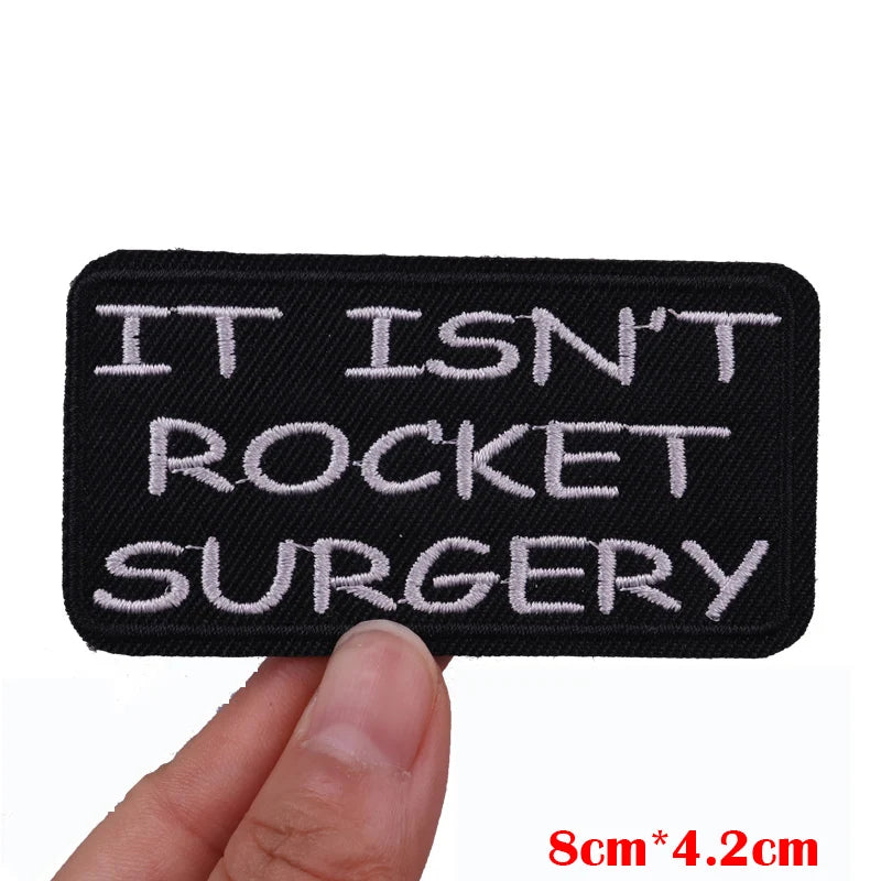 Black embroidered patch with white text reading IT ISN'T ROCKET SURGERY being held by a hand