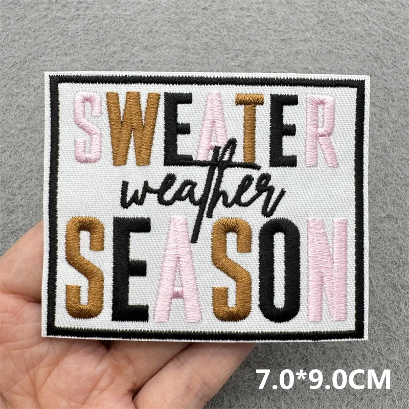 Black and pink embroidered text on a white patch that reads SWEATER WEATHER SEASON