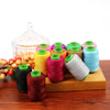 A collection of 24 spools of colored sewing threads in various shades of green, pink, yellow, and blue resting on a wooden bo