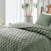 A close-up of a sage green quilted duvet and pillow set on a bed, with a potted plant in the background.
