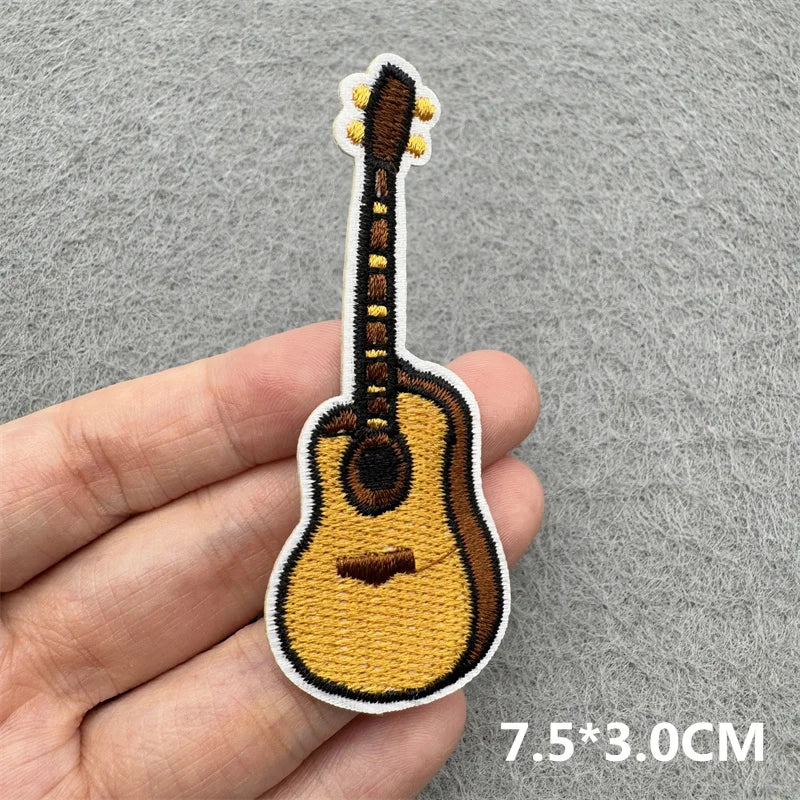 An embroidered patch of a brown acoustic guitar with a yellow body and white pickguard, shown being held by a hand.