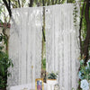 A sheer white floral lace curtain panel hanging outdoors in a garden setting.
