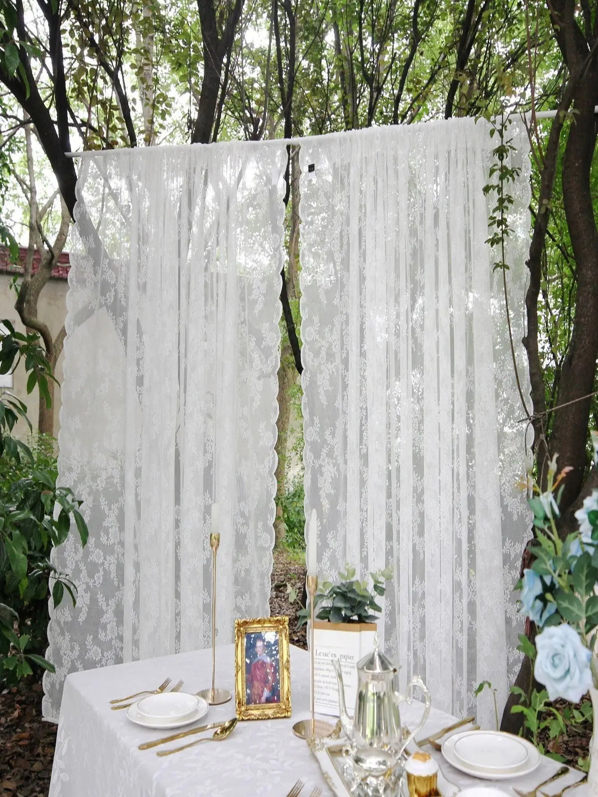 A sheer white floral lace curtain panel hanging outdoors in a garden setting.
