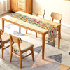 A light wood dining table set with matching chairs and a colorful Day of the Dead linen table runner on a patterned rug.