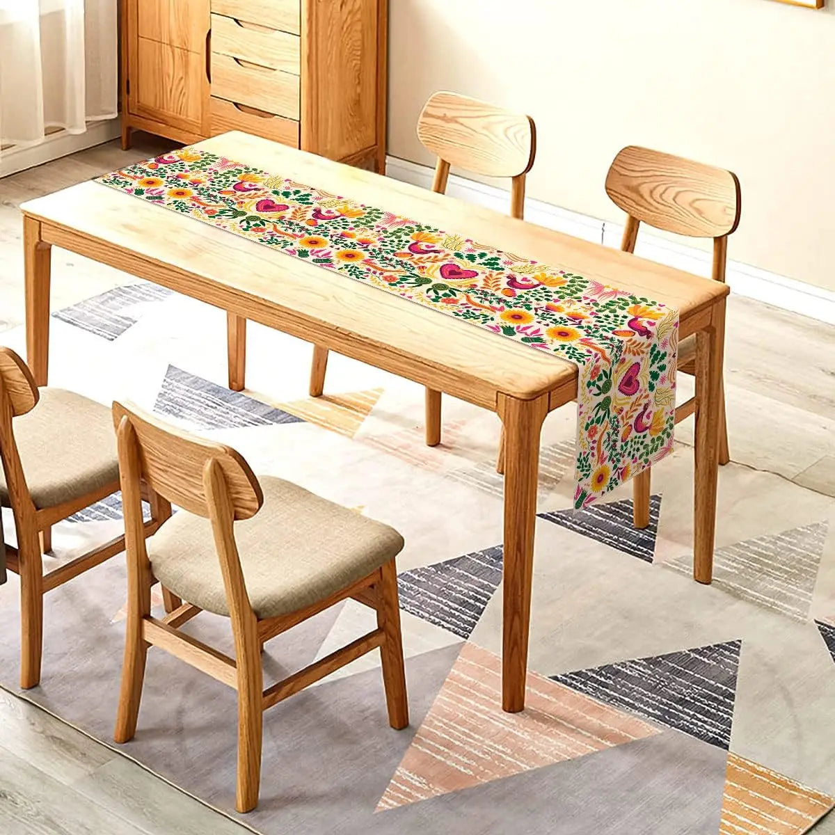 A light wood dining table set with matching chairs and a colorful Day of the Dead linen table runner on a patterned rug.