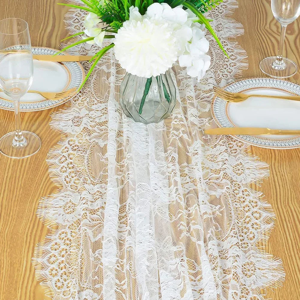 A white embroidered floral lace table runner laid on a wooden table with a vase of white flowers and place settings.