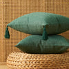 A deep green linen throw pillow with tasseled trim rests on a woven natural fiber surface against a textured wall.